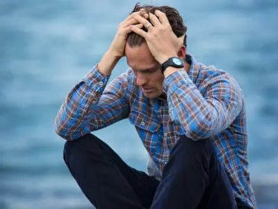 Man sits on a rock by the ocean with his heads on his head looking stressed.