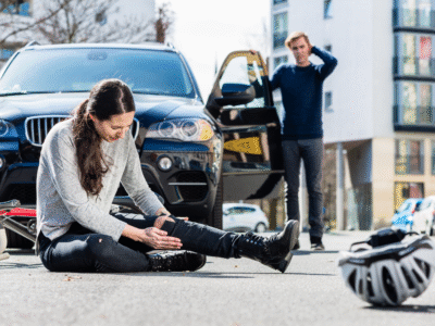 A bicyclist on the street holding their leg and a car is stopped behind them the driver is stressed.