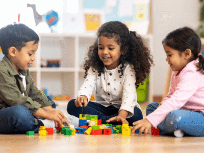 Three young children sit on the floor in a brightly lit classroom, smiling and playing with colorful building blocks, symbolizing the legal challenges Atlanta day care abuse attorneys face when handling child injury and abuse cases.