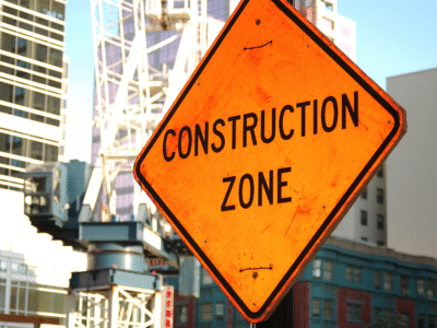 Bright orange “Construction Zone” warning sign stands in front of high-rise buildings and cranes, representing third-party construction injury claims in Georgia involving subcontractor liability, contractor negligence, and road construction accidents.