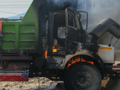 A commercial truck with a severely burned cab sits on the roadside, with visible flames still flickering inside and around the wheel well as smoke rises into the air. The damage suggests a catastrophic incident, potentially caused by mechanical failure, poor maintenance, or a serious collision—factors often central to wrongful death and negligence claims involving large trucks.