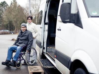 Elderly man in a wheelchair being assisted out of a transport van by a caregiver, illustrating potential safety concerns and liability in nursing home transportation accidents.